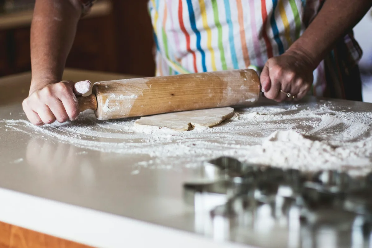 A home baker in a rainbow-striped apron rolling out cookie dough with a wooden rolling pin on a flour-dusted kitchen counter, with metal cookie cutters waiting in the foreground