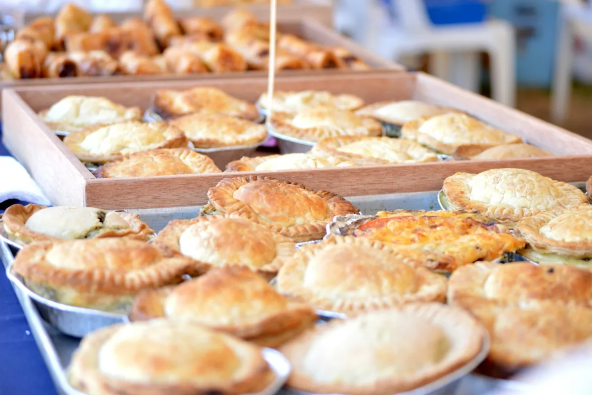 Trays of golden-brown meat pies and empanadas plus a quiche arranged on a market table, with additional pastries on a wooden tray in the background, photographed in warm natural light at an outdoor farmers market