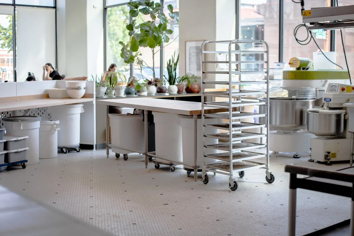 A bakery workroom with a loaded sheet-pan rack and dough tubs beside a butcher-block worktable