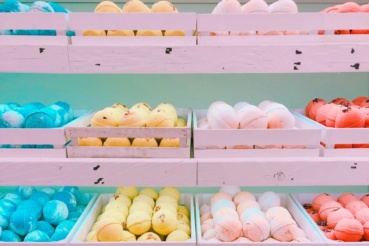 Crates of colorful handmade bath bombs in blue, yellow, peach, and coral arranged on white shelves in a shop display