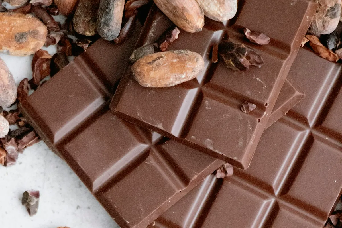 Stacked bars of dark chocolate surrounded by whole roasted cacao beans and scattered cocoa nibs on a pale surface