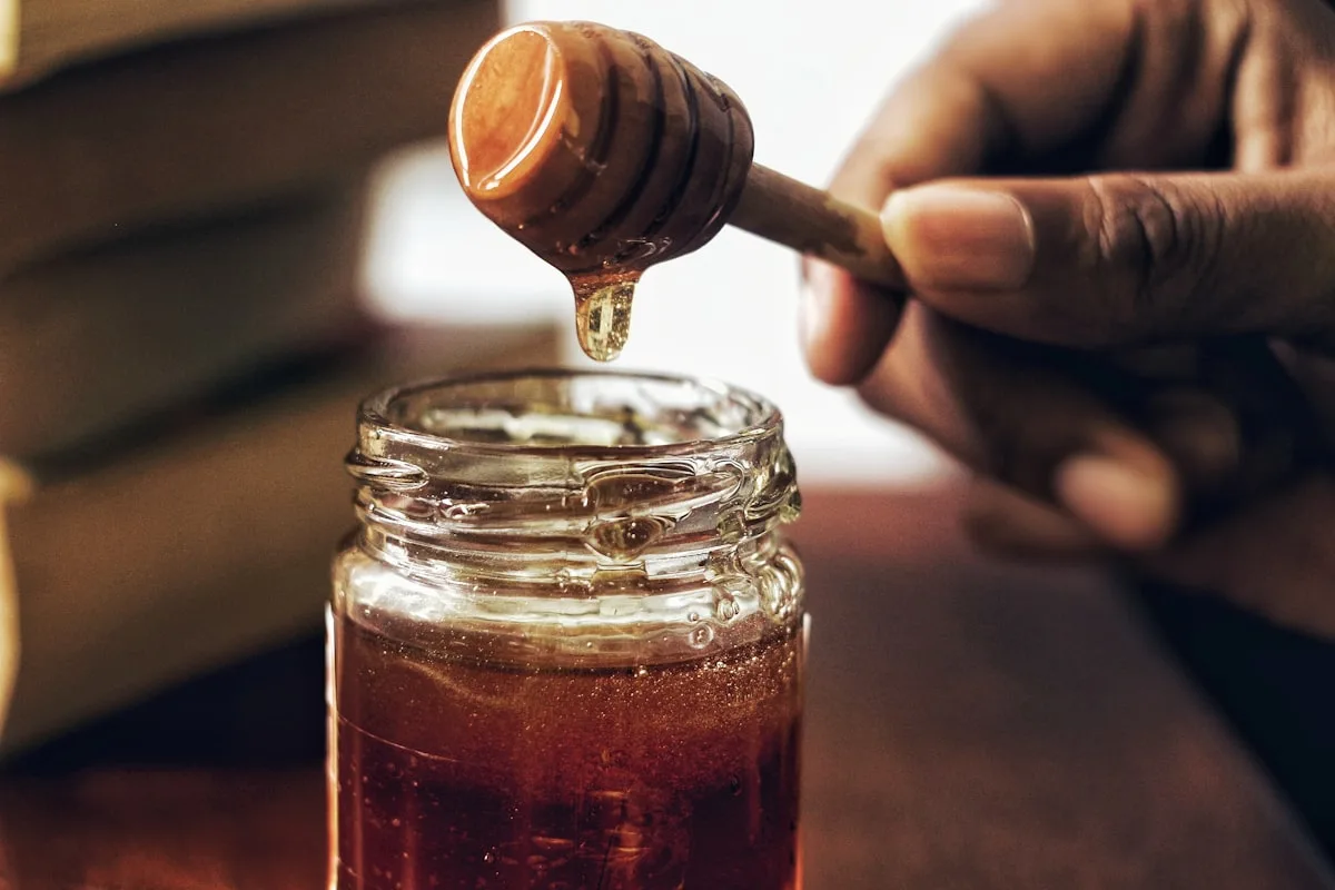 A wooden honey dipper drizzling harvested honey into a clean glass jar during the bottling step