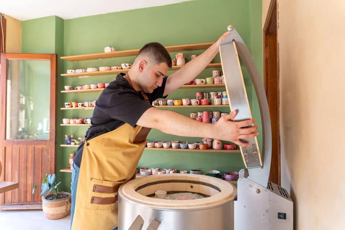 A potter in a mustard apron lifts the hinged lid of a round stainless-steel electric kiln, in front of a green wall lined with shelves of hand-painted mugs and cups