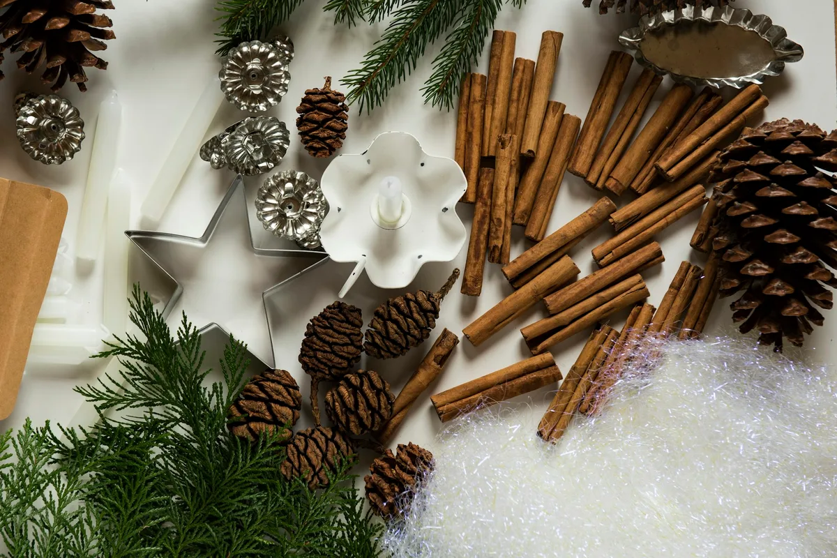 Overhead view of holiday craft supplies on a white table: cinnamon sticks, pine cones, evergreen sprigs, white taper candles, a star cookie cutter and silver candle clips beside a drift of white angel hair
