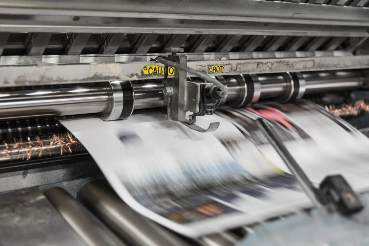 Printed sheets running at speed through the steel rollers of a large offset printing press, the pages blurred with motion