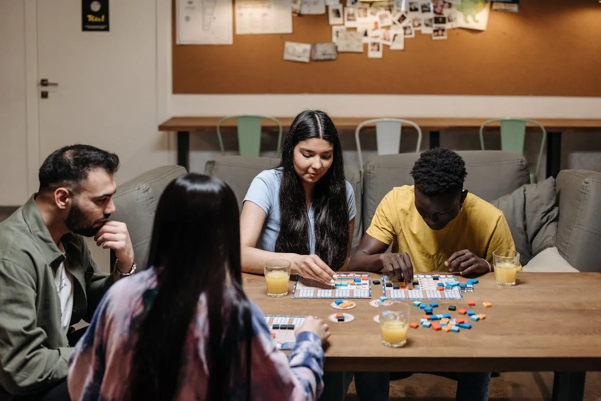 A group of friends playing a colorful board game with tiles and game pieces spread across a wooden table in a casual lounge setting