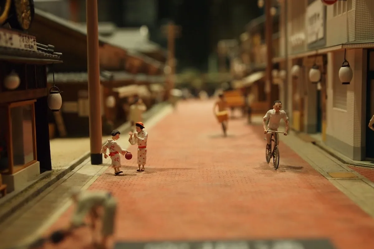 A finely detailed miniature street-scene diorama — tiny figures in kimono, a cyclist, and hanging paper lanterns along a model town alley.