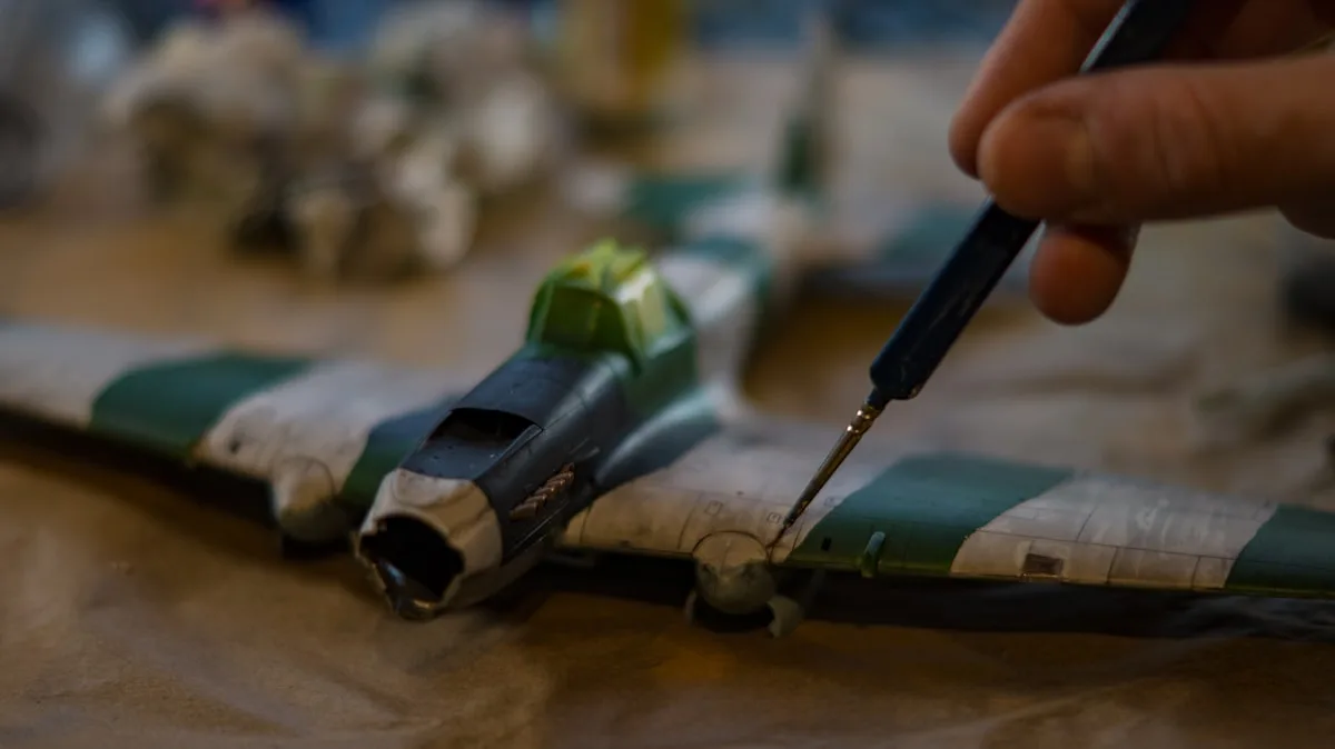 A hobbyist's hand steadying a fine tool to detail a small painted model on a workbench — the painstaking assembly behind a finished miniature.
