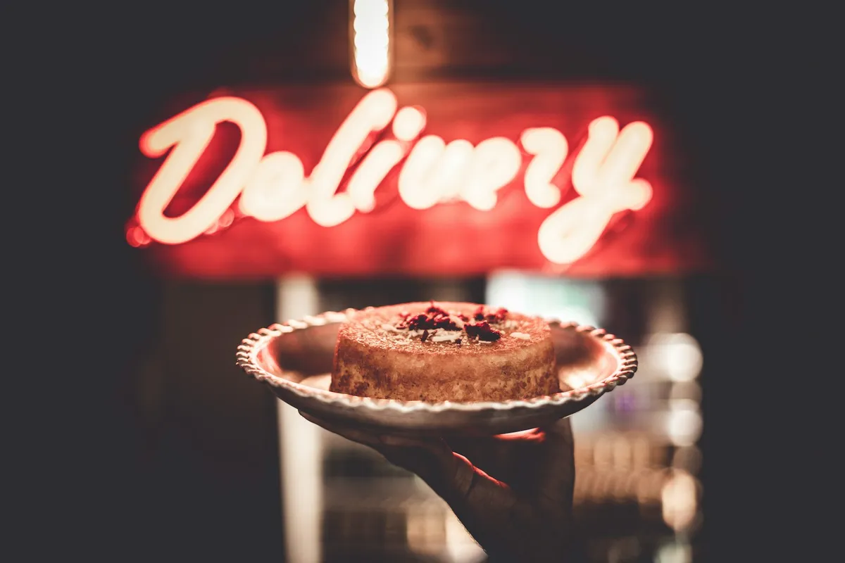 A hand holding a small cake on a scalloped plate in front of a glowing red neon sign that reads Delivery