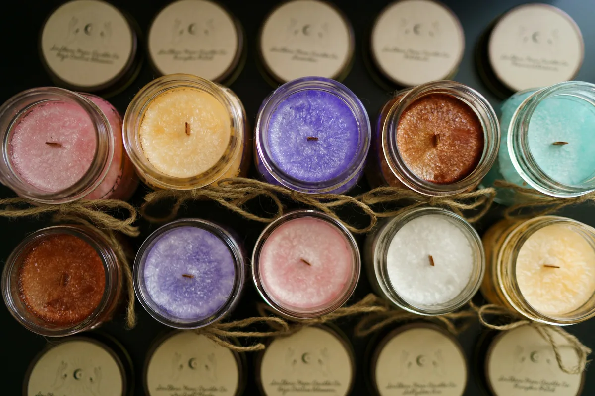 Overhead view of ten unlit handmade candles in pink, yellow, purple, brown, teal and white wax, their jars uncapped and tied with twine, with the lids set out in rows above and below