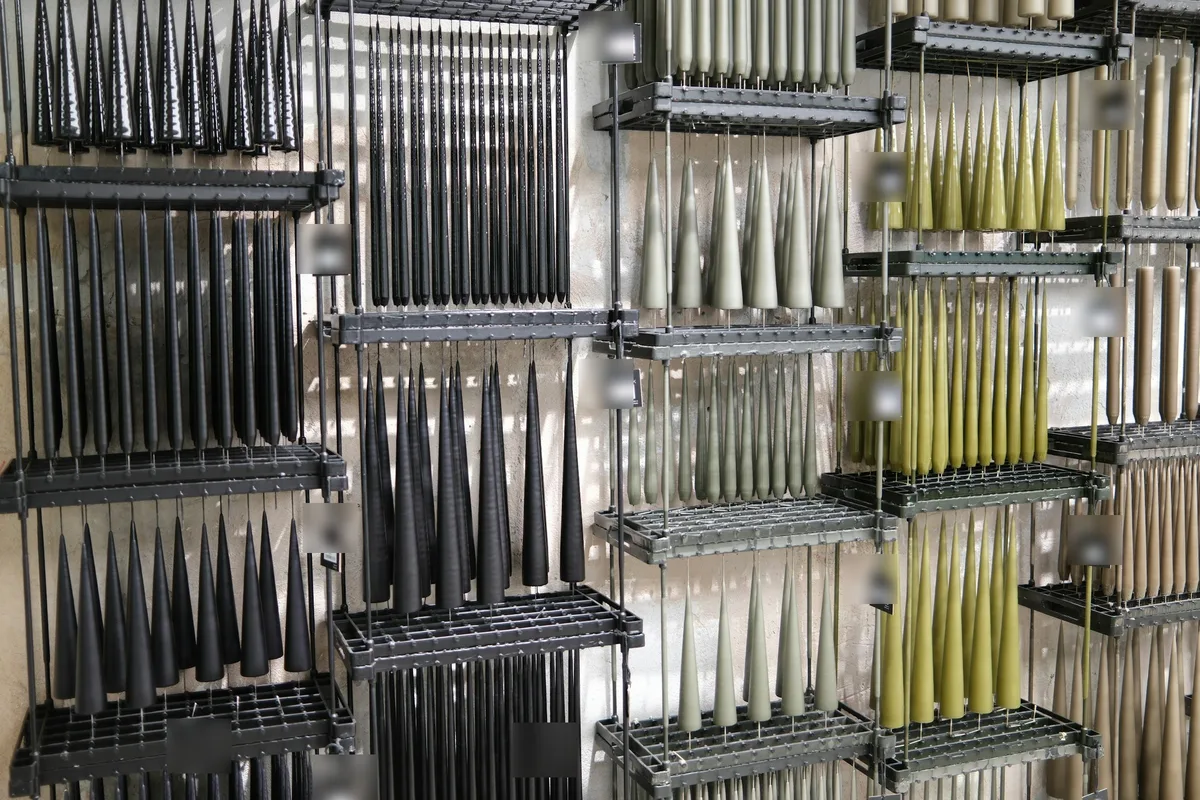 Tiered metal drying racks holding dozens of tapered candles in four colorways — black, sage, olive, and sand — hanging in rows against a plaster wall