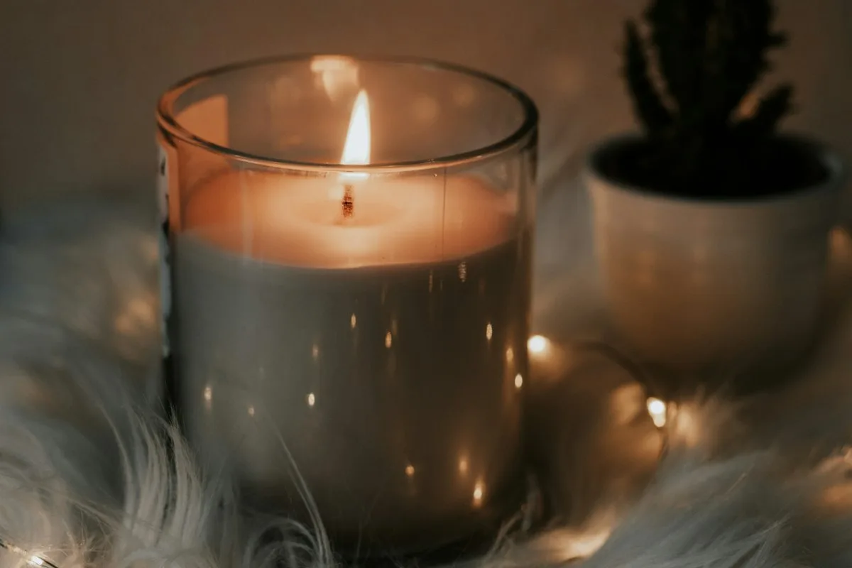 A lit candle in a clear glass jar resting on a soft white throw, its flame steady above a shallow melt pool, with fairy lights and a small potted plant blurred behind it
