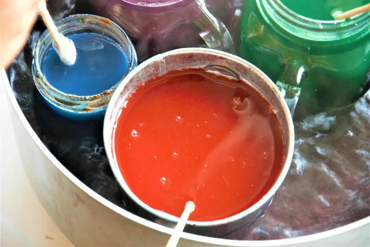 A metal tin of melted red wax and glass jars of blue, purple and green wax standing in a hot water bath inside a large pot, with wooden stir sticks resting in the wax
