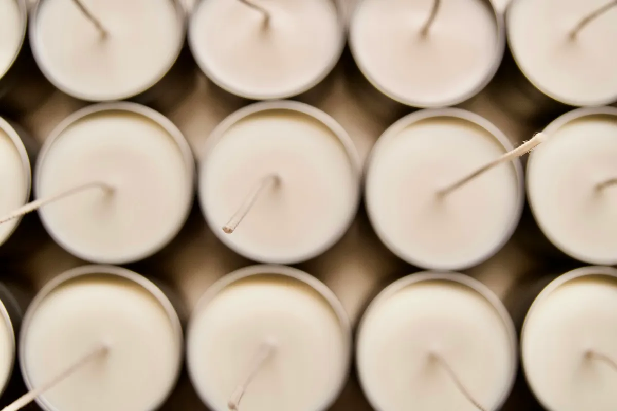 Rows of freshly poured white container candles seen from directly above, each with an untrimmed cotton wick leaning off center