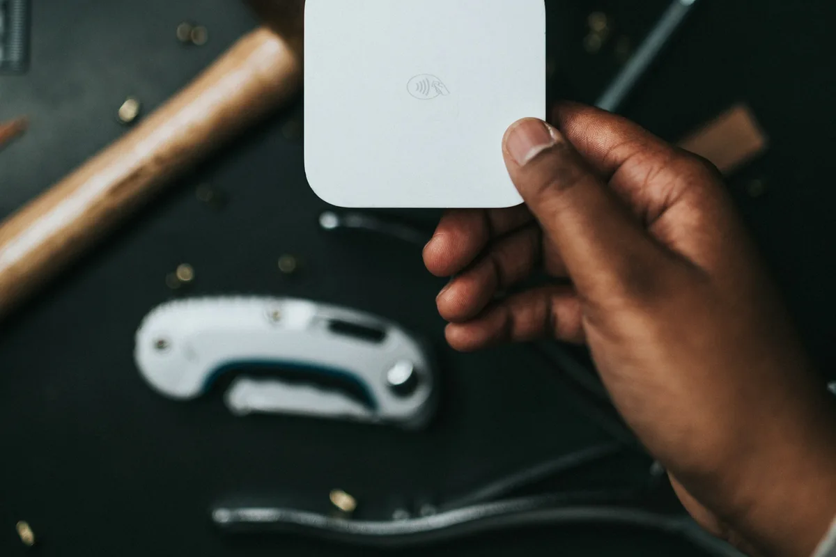 A hand holding a small white contactless card reader above a leatherworker's bench, with a wooden mallet handle, a folding utility knife and scattered brass rivets on the dark work surface below