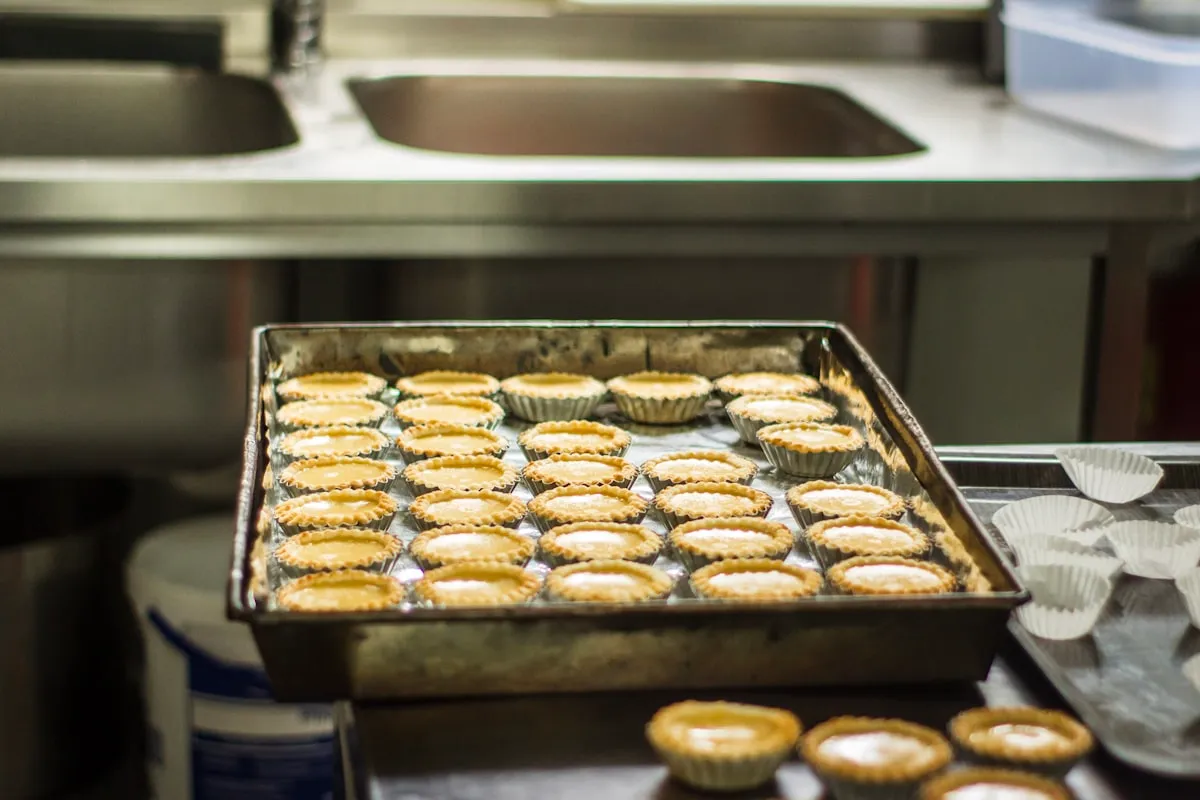 A full sheet pan of small tart shells cooling on a stainless prep table in a commercial kitchen, with a double sink and a stack of empty paper cases behind it