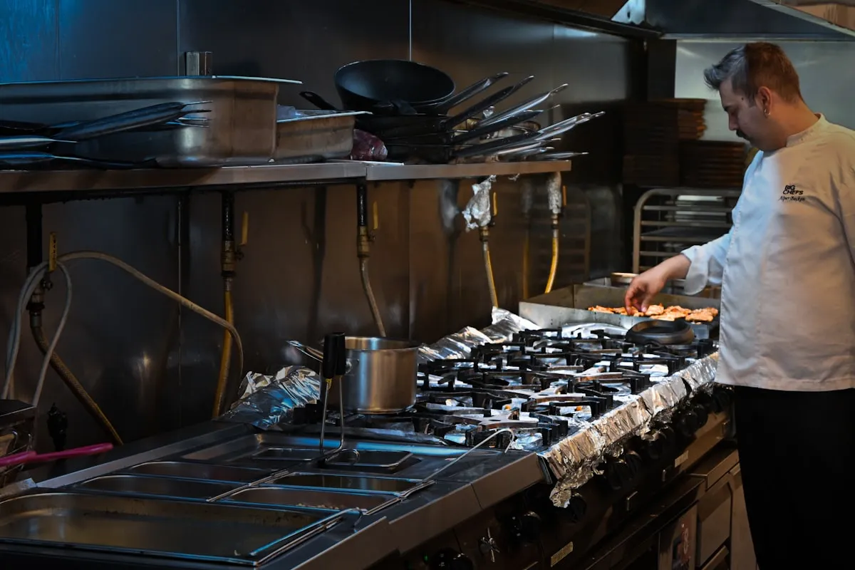 A cook in whites working alone at a stainless commercial range line, foil-lined burners in front and stacked pans and sheet trays on the shelf above