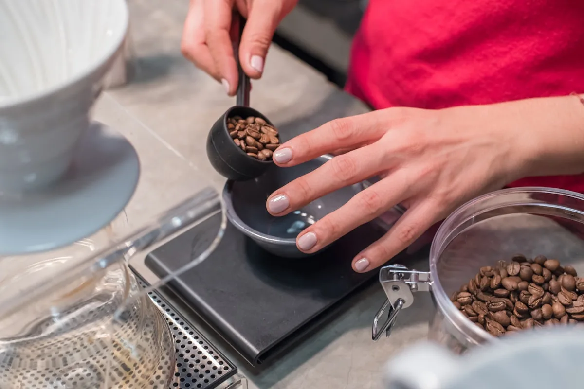 Hands tipping roasted coffee beans from a scoop onto a flat digital scale platform, with a jar of beans alongside on the counter