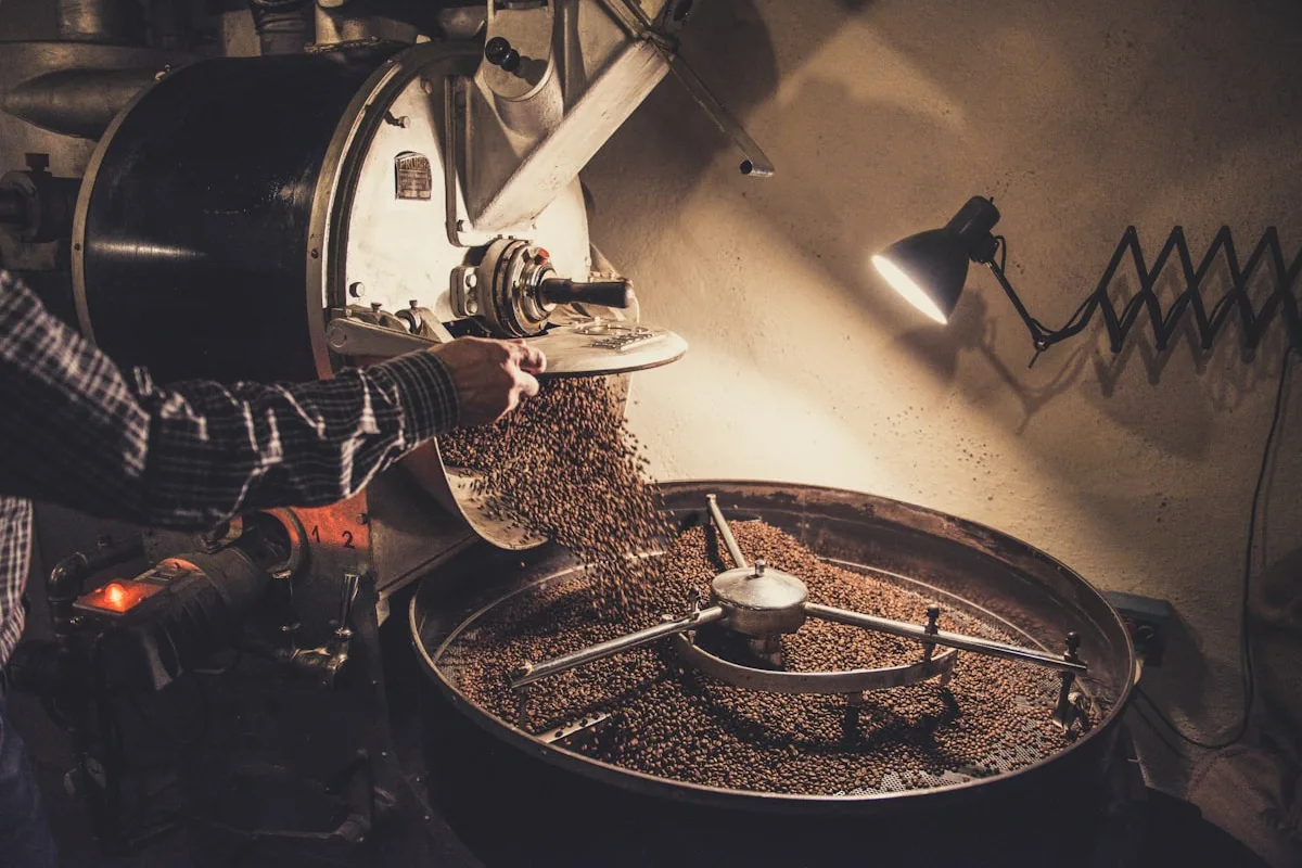 A coffee roaster tipping freshly roasted beans from the drum into a spinning cooling tray, a worker's arm guiding the chute under a warm work lamp