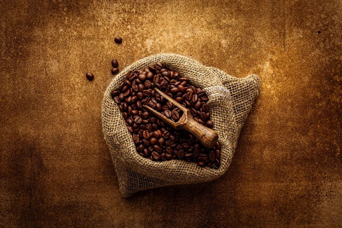 Roasted coffee beans in an open burlap sack with a wooden scoop, viewed from above on a dark textured surface