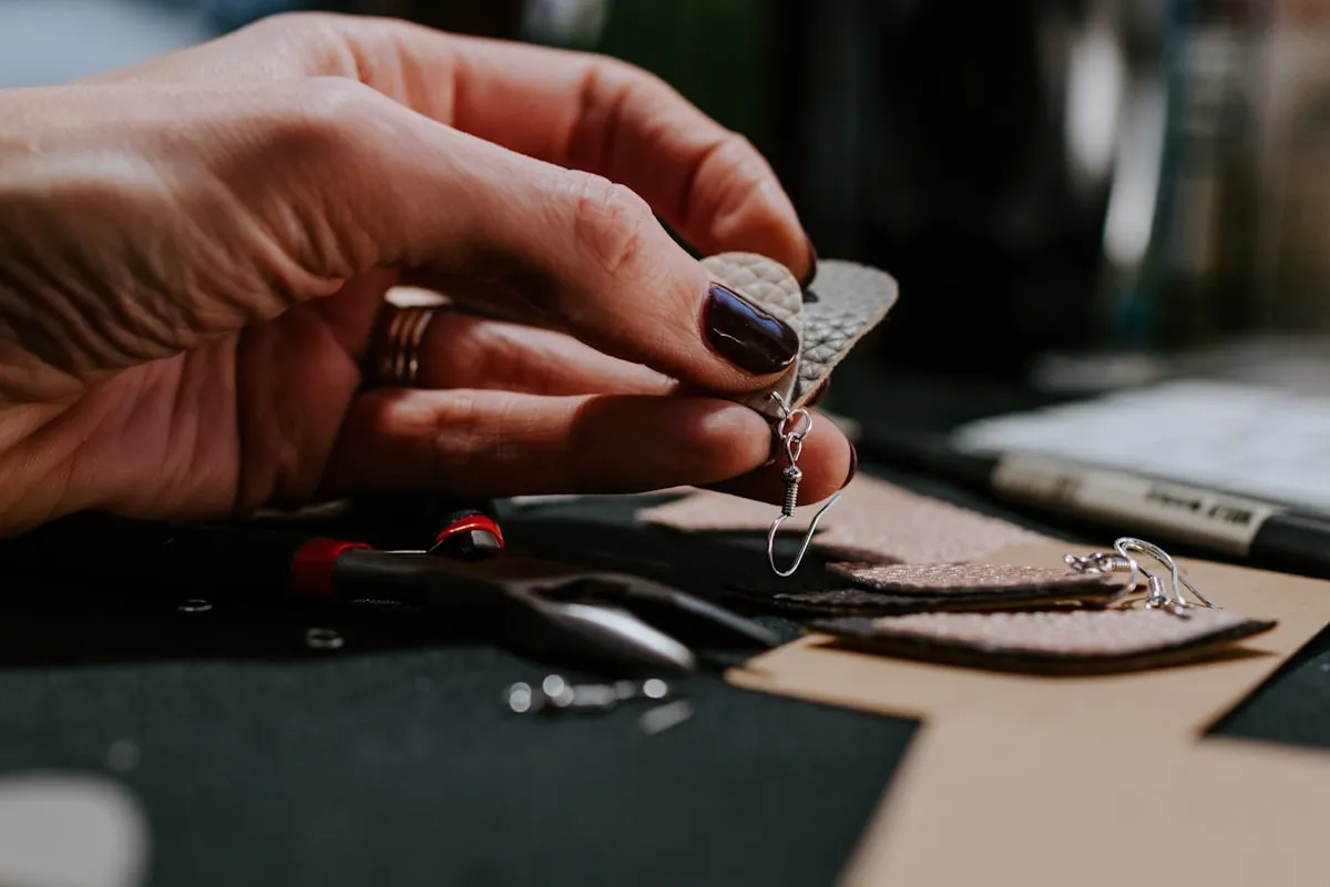 A maker's hand assembles a small leather earring with a silver finding on a workbench surrounded by leather scraps, hardware, and pliers in warm afternoon light