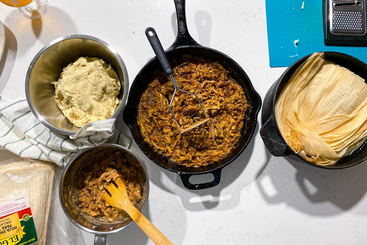 An overhead view of a home kitchen counter mid-production: a cast-iron pan of shredded meat filling, a bowl of yellow masa dough, a pot of soaked corn husks, and a saucepan of seasoned filling beside a folded dish towel — the kind of home-kitchen tamale production the Colorado Tamale Act (HB26-1033) authorizes for cottage food sales for the first time