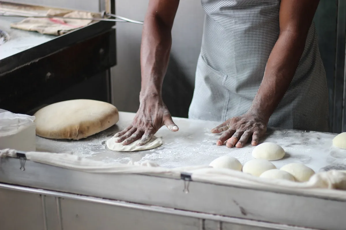 A baker in a checkered apron presses and shapes dough on a flour-dusted work surface, with a finished round loaf and several pre-shaped dough balls resting on linen cloth nearby