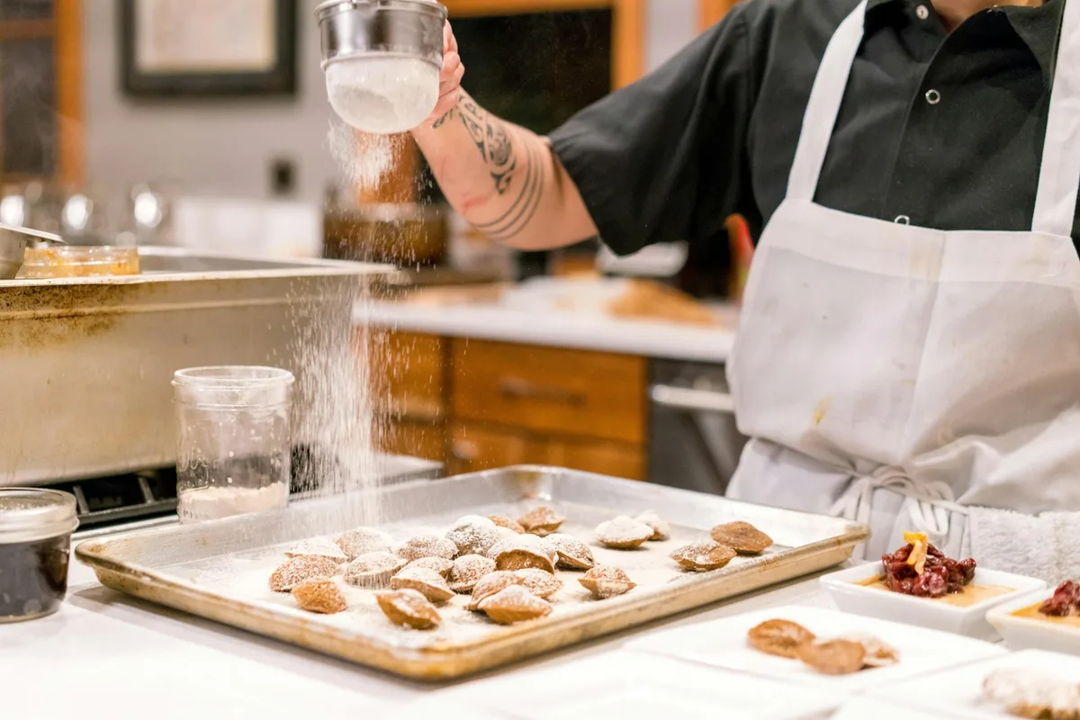 An aproned baker dusting powdered sugar over a tray of freshly baked pastries on a home kitchen counter