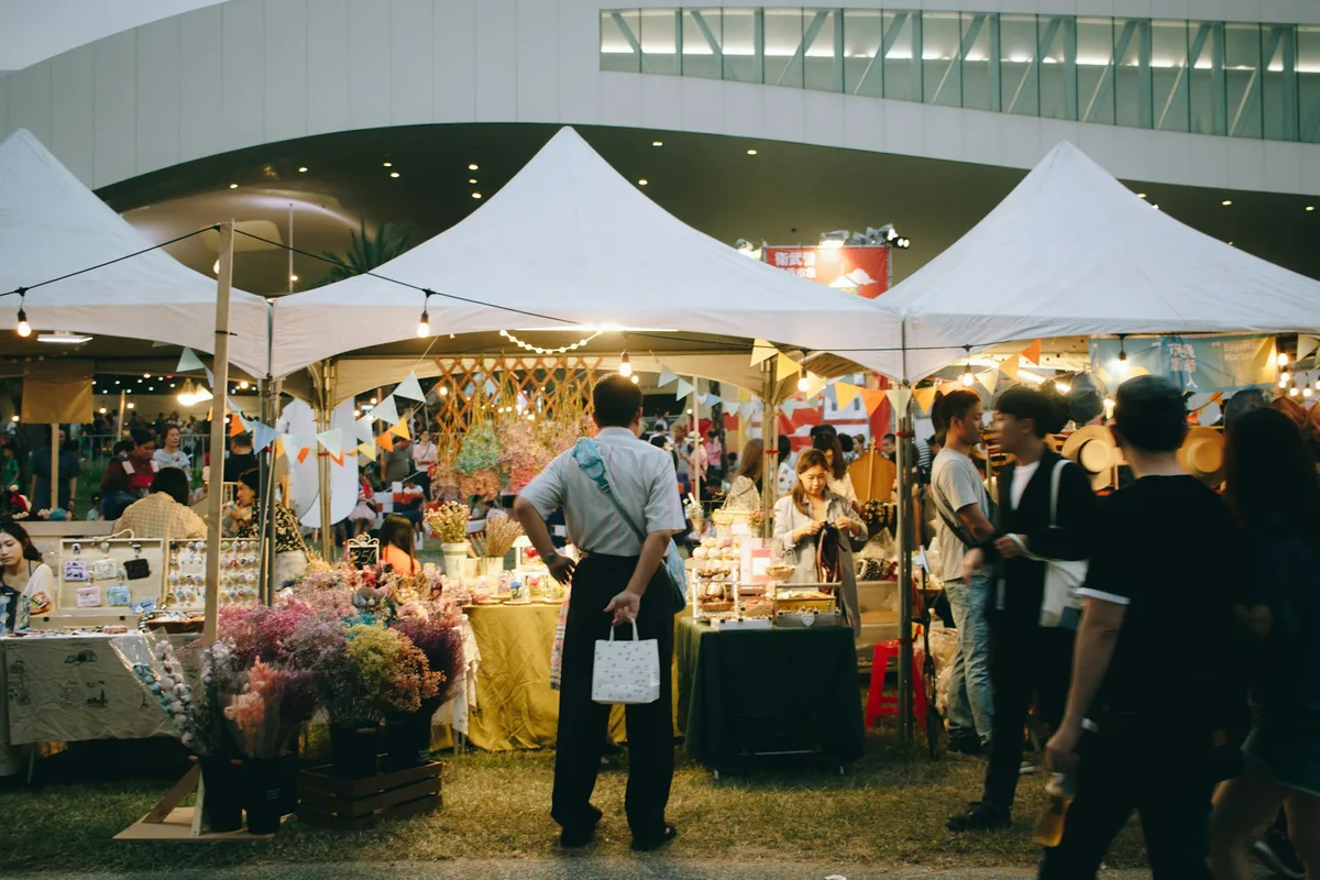 An evening craft market with white canopy tents strung with lights, tables of handmade goods and dried flowers, and shoppers browsing between the booths