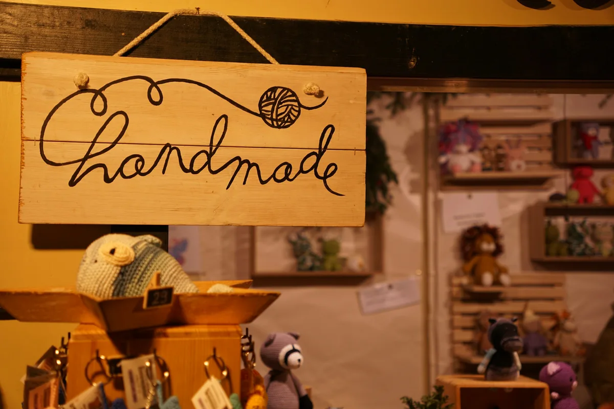 A hand-lettered wooden sign reading handmade above a craft market stall of crocheted animals and keyrings on wooden shelves