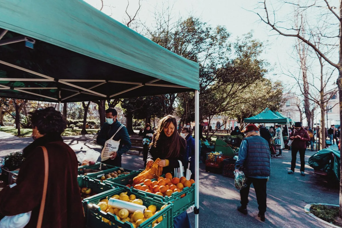 A teal pop-up canopy over an outdoor market stall, its truss frame and straight white leg visible above crates of oranges and lemons, with a second canopy further down the paved row and shoppers walking past