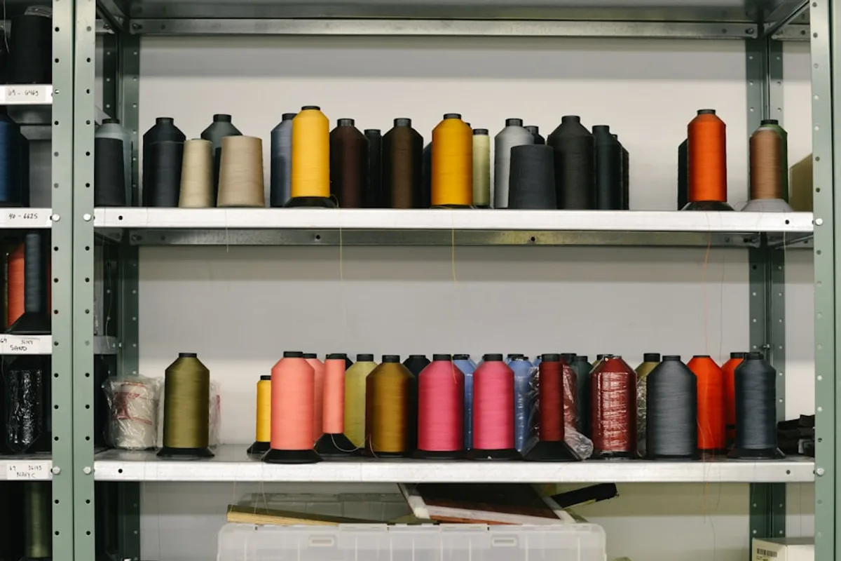 Industrial thread cones in black, mustard, coral and blue on steel shelving, with handwritten inventory labels along the shelf edges