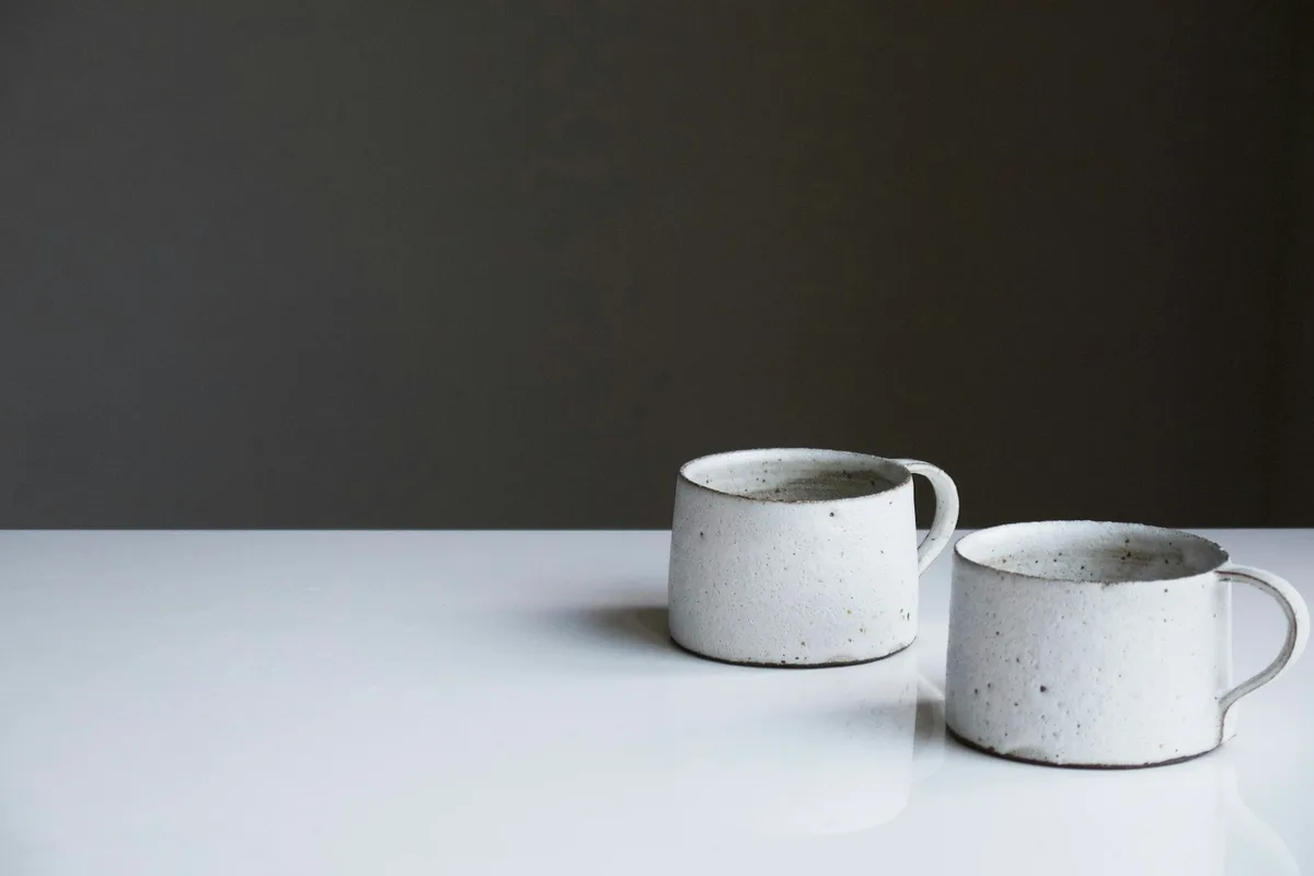 Two handmade speckled white ceramic mugs sitting side by side on a glossy white tabletop against a soft charcoal-gray wall, lit from the right