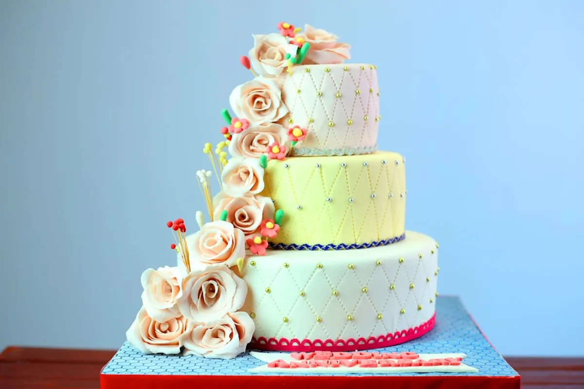 Three-tier fondant celebration cake with cascading sugar roses and small pink blossoms on a blue patterned cake board against a soft blue background