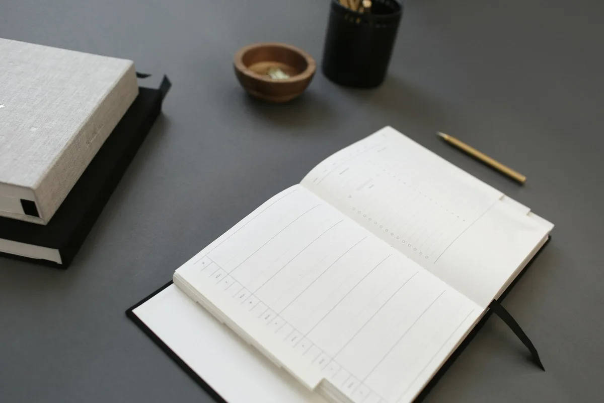 An open ruled order ledger and a slim brass pencil on a gray desk, beside two closed notebooks and a small wooden bowl