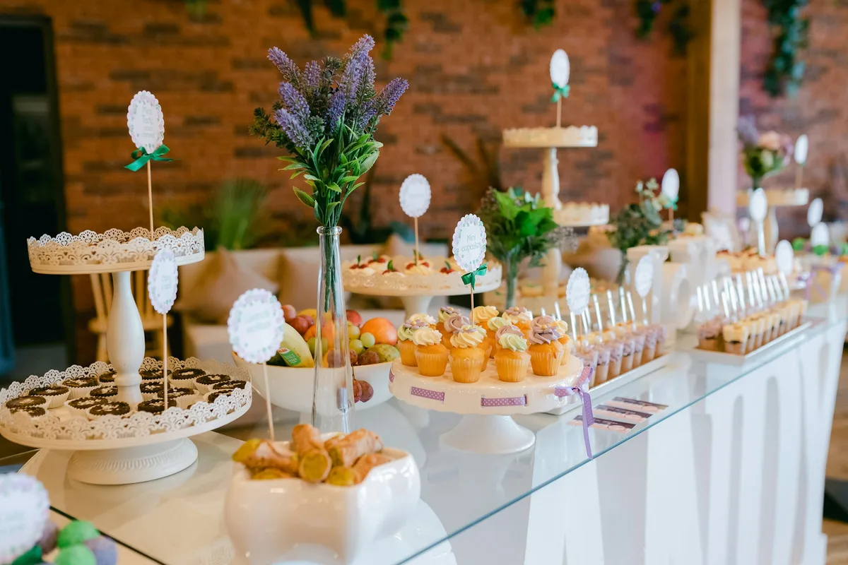 A long glass-topped event dessert table against a brick wall, set with white lace cake stands, mini cupcakes with pastel frosting, rows of dessert cups and small paper label flags beside a vase of lavender
