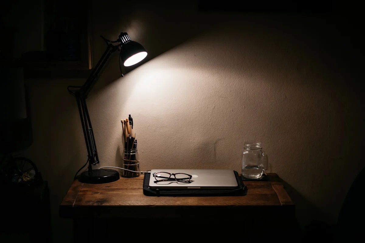 A closed laptop, reading glasses and a jar of pencils on a wooden desk in a dark room, lit by one angled desk lamp