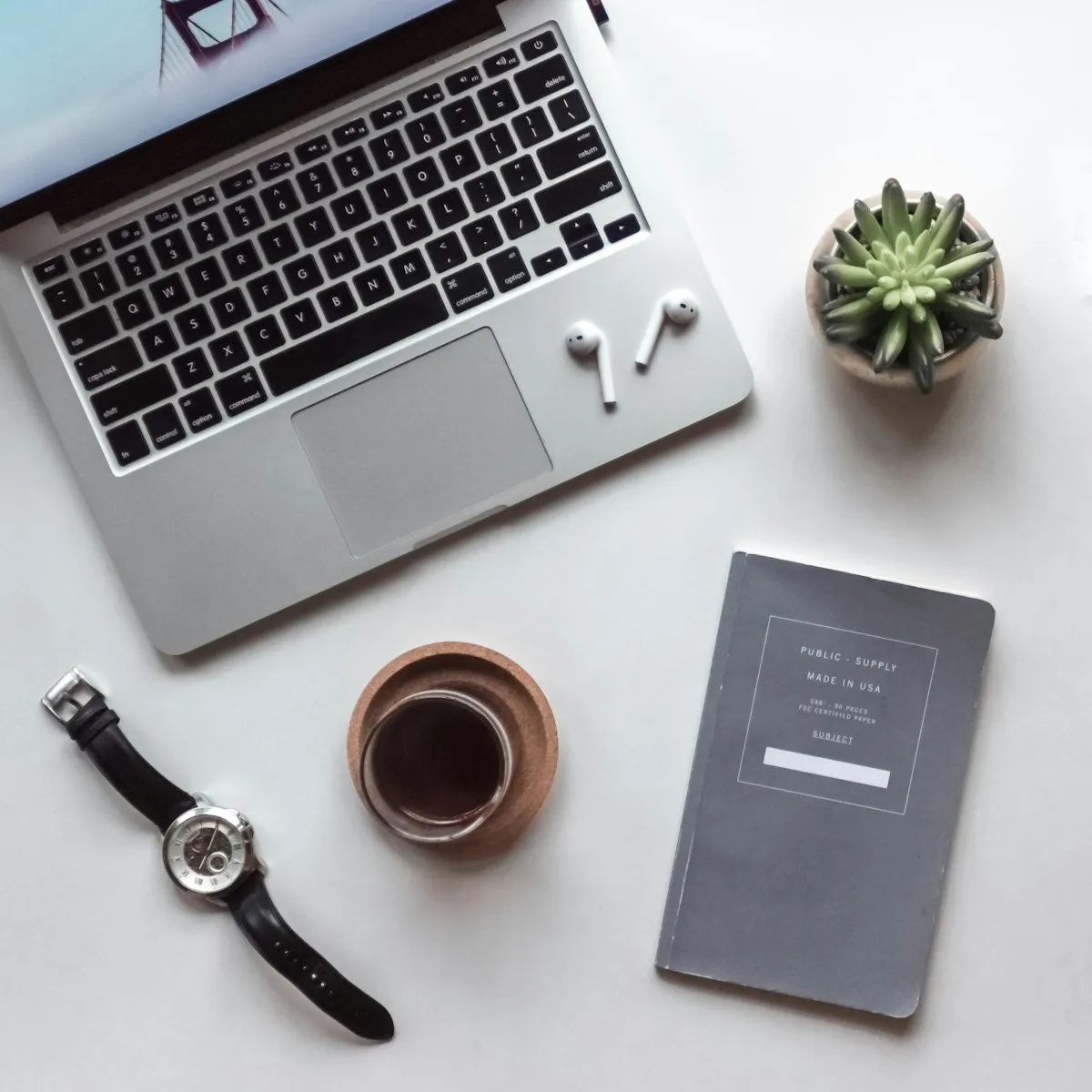 Flat lay of a MacBook laptop with coffee, notebook, watch, earbuds, and a small succulent on a clean white desk