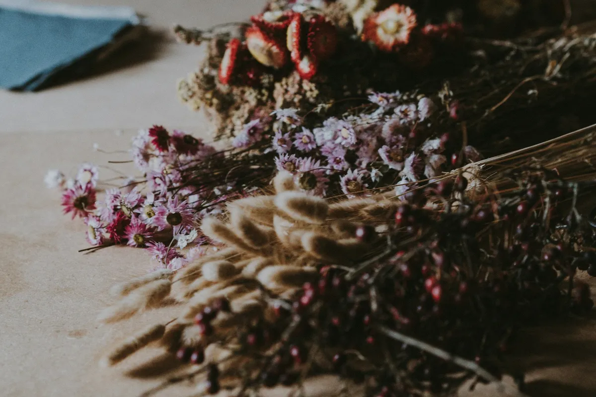 Dried flower bunches laid out on a kraft-paper work table ready for assembly, including red and white strawflowers, pink and lilac everlastings, pale bunny-tail grass, and sprigs of dark berries