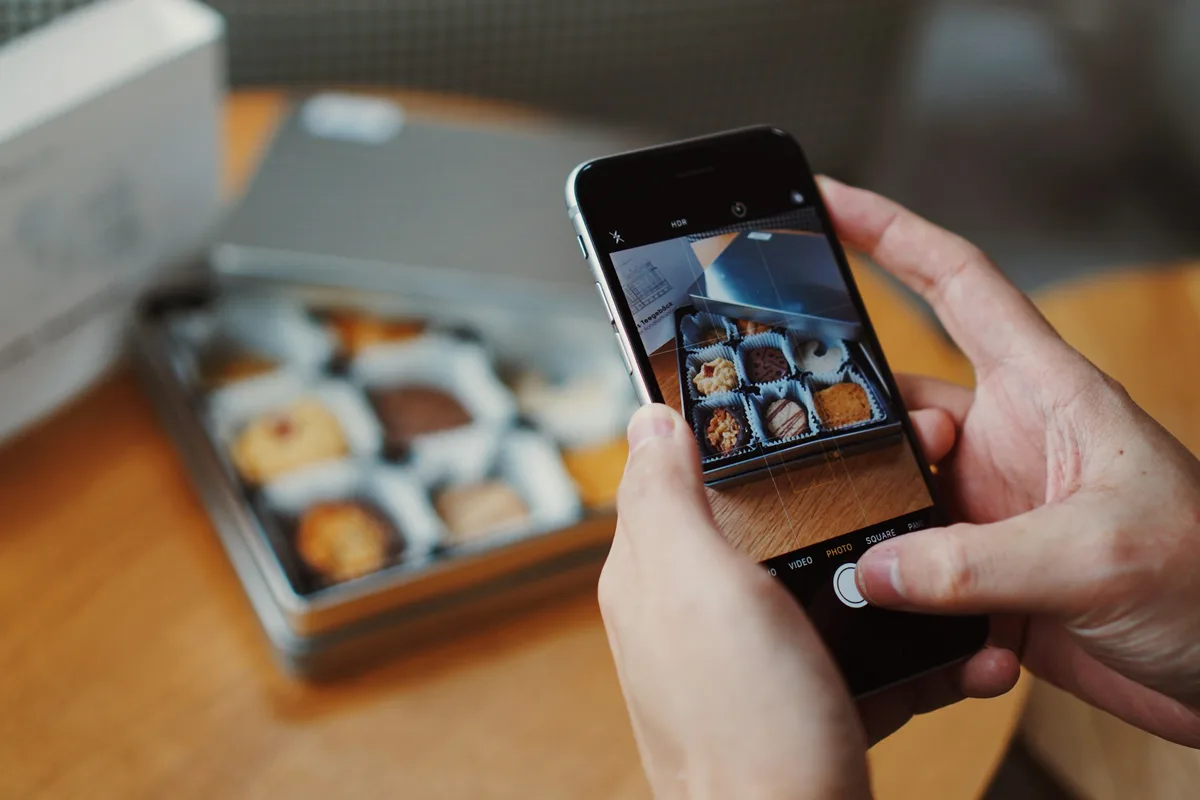 Two hands hold a phone photographing an open tin of assorted cookies in paper cups on a wooden table, the phone screen showing the same box in natural light