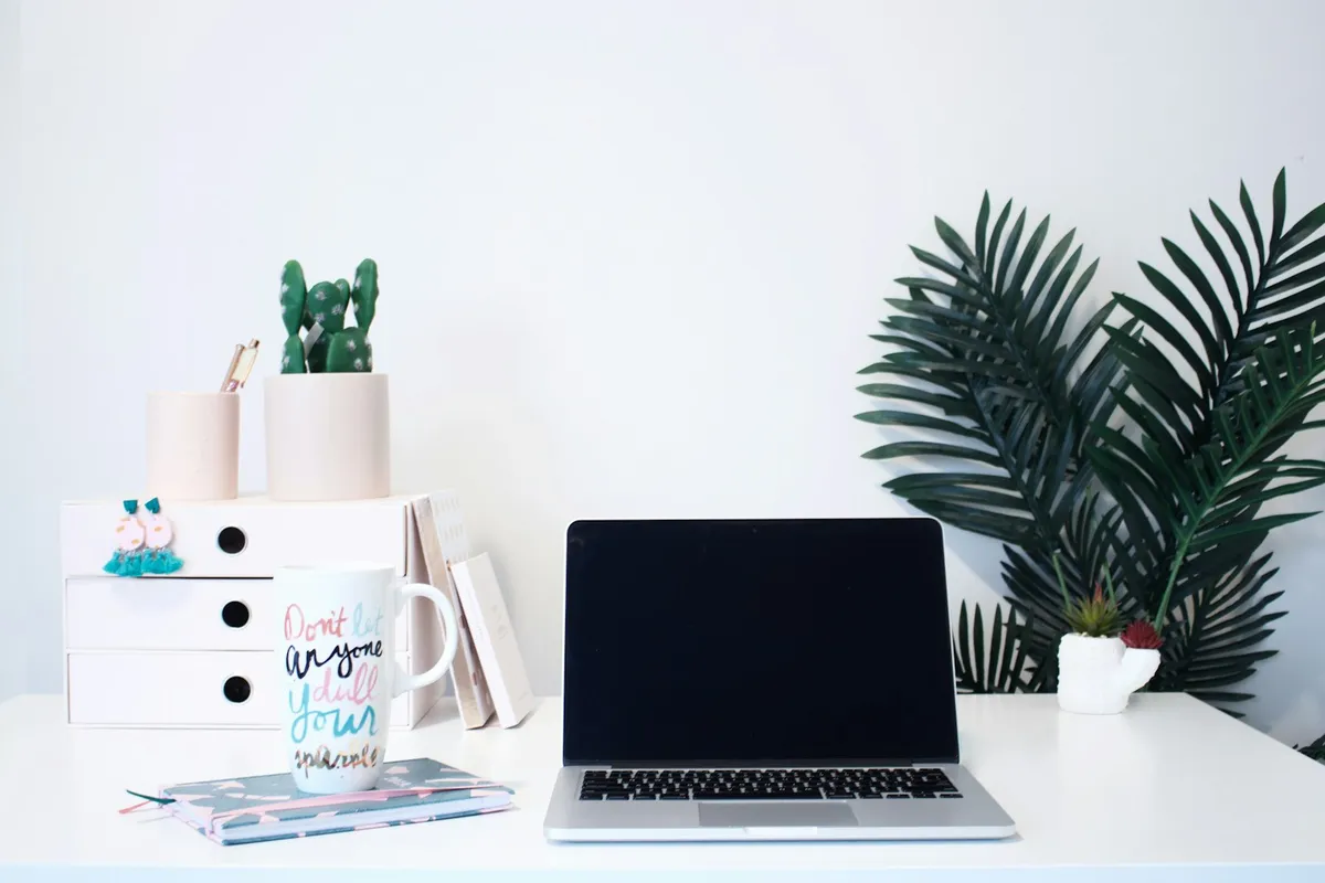 A tidy white desk with an open silver laptop, a stack of three white organizer drawers holding pastel ceramic cups, a small cactus planter, a pen, a tasseled handmade earring hung on the drawer pull, a hand-lettered "Don't let anyone dull your sparkle" mug resting on a teal patterned notebook, and a tall palm and small succulent against a soft white wall