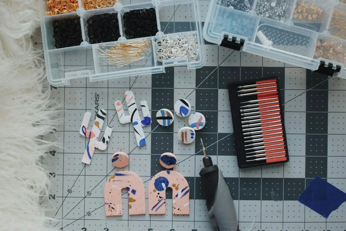 Overhead view of a cutting mat with pairs of hand-painted polymer clay earrings, including pink arch shapes, below two clear organizer boxes full of jump rings, eye pins and other earring findings, with a rotary tool and a tray of bits beside them