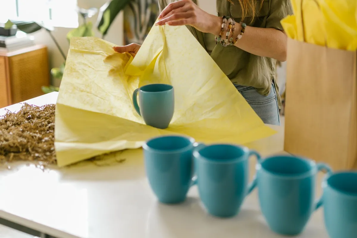 A maker wraps a teal ceramic mug in yellow tissue paper to ship an order, with four matching mugs lined up in front