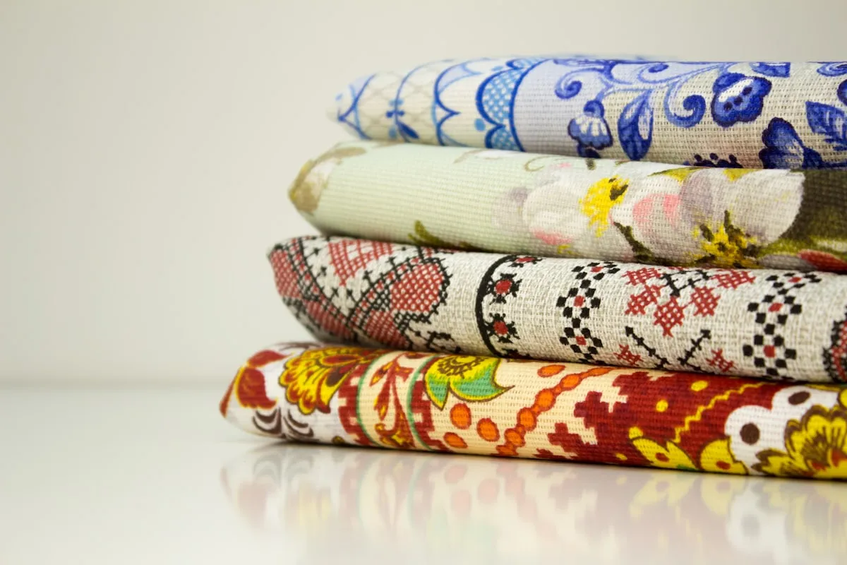 A stack of four folded printed cotton fabrics on a white surface — a blue floral, a pale green botanical, a red and black geometric, and a red and yellow folk print