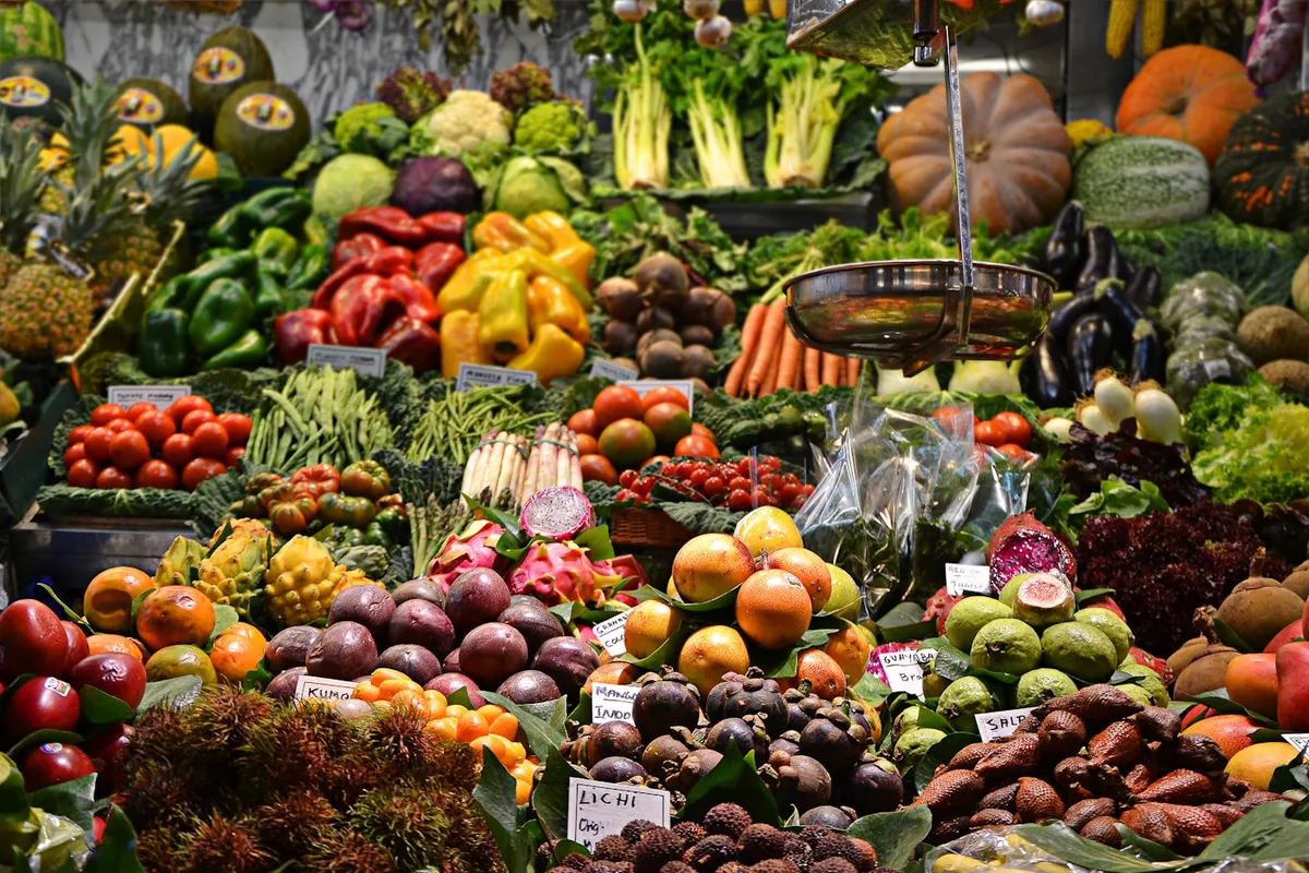 A market stall piled high with colorful fruit and vegetables — tomatoes, peppers, citrus, squash, and tropical fruit in crates with small handwritten product labels