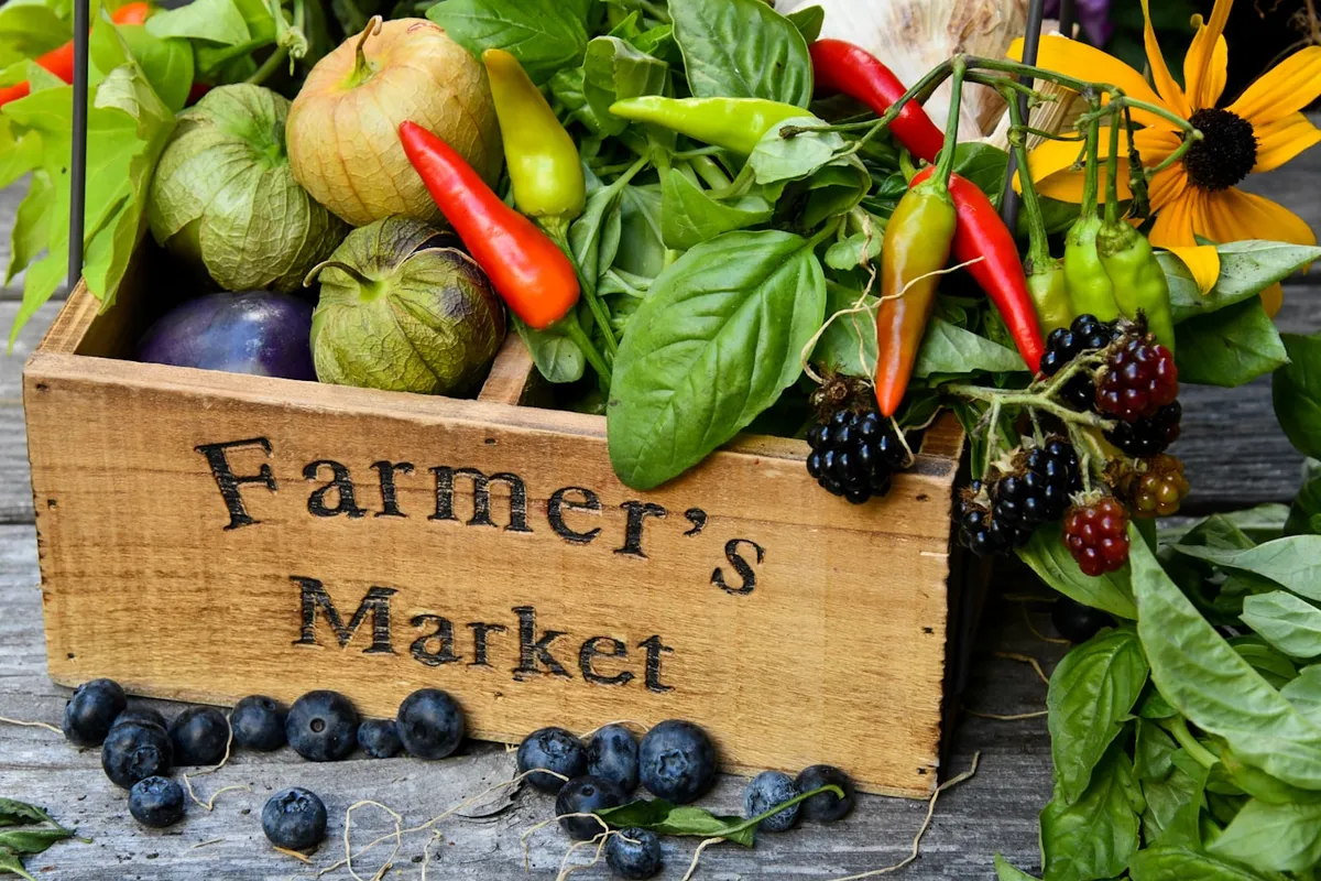 A wooden crate stamped "Farmer's Market", filled with peppers, tomatillos, basil, and blackberries, with blueberries and a sunflower on weathered wood