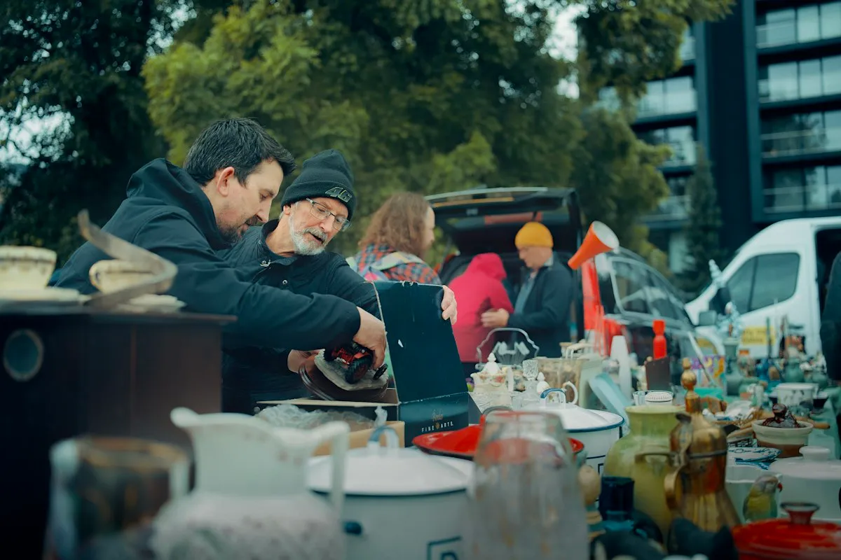 Two shoppers, one in a black knit hat, lean over an outdoor market table crowded with vintage jugs, enamel pots and glassware, lifting a small item out of an open box while other visitors browse behind them