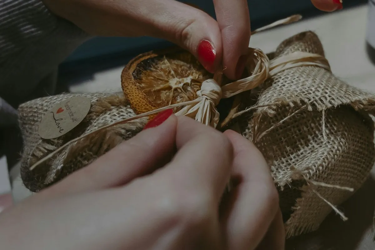 Close-up of two hands wrapping a small handmade parcel — burlap fabric tied with raffia twine, decorated with a dried orange slice and a small kraft tag, against a softly lit dark wooden surface