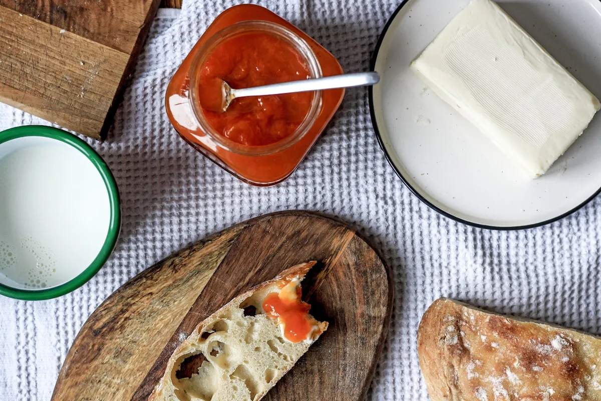 An overhead flat lay on a textured white linen with an open glass jar of orange apricot preserves and spoon, a wedge of butter on a small plate, an enamel cup of milk, and a torn slice of crusty sourdough resting on a wooden cutting board next to the rest of the loaf