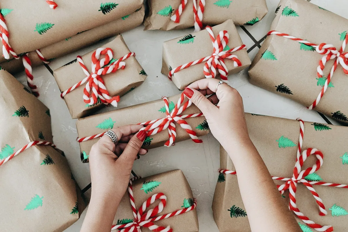 Two hands tying red-and-white twine around a kraft-paper gift printed with small green trees, surrounded by more boxes wrapped and tied the same way
